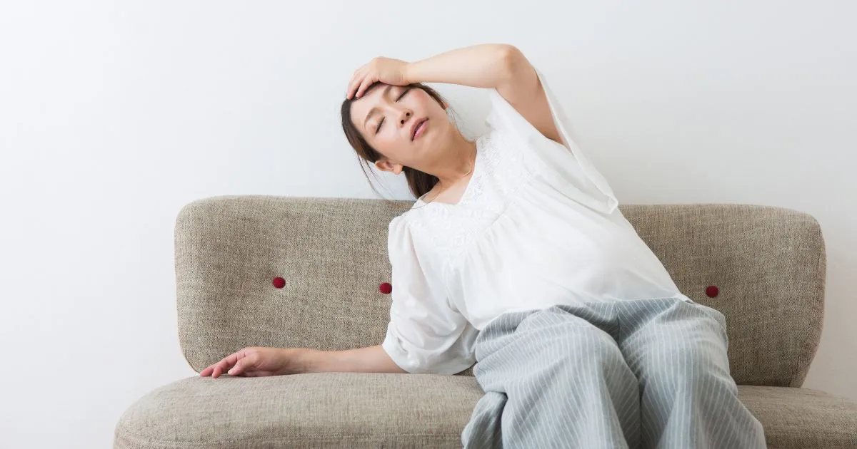 ソファに座り頭を押さえながら不安定さを感じている女性(めまいのイメージ)